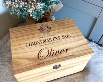 May include: A rectangular wooden box with a hinged lid and a metal latch. The lid is engraved with a crest, the text SPECIAL DELIVERY CHRISTMAS EVE BOX, FOR THE ATTENTION OF: Oliver, and PROPERTY OF SANTA CLAUS, NORTH POLE in dark lettering.