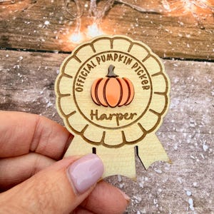 Peut inclure: Un badge en bois en forme de ruban de récompense circulaire avec un bord festonné, gravé du texte OFFICIAL PUMPKIN PICKER et du prénom Harper. Un graphique de citrouille orange est centré sur la surface en bois clair du badge.