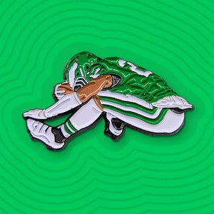 May include: Enamel pin depicting a football player in a green and white uniform, head in hands, in a dejected pose. The pin is set against a green background with a layered, topographic design.