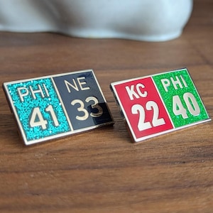 May include: Two silver pins with a black, red, and green color scheme. The pins show the scores of two football games. The first pin shows "PHI 41 NE 33" and the second pin shows "KC 22 PHI 40".
