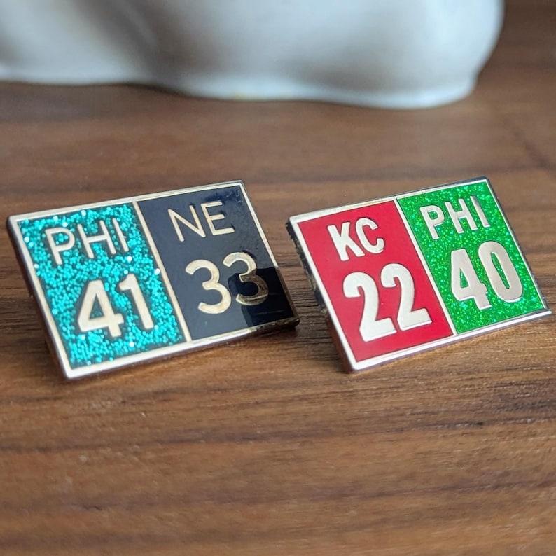May include: Two silver pins with a black, red, and green color scheme. The pins show the scores of two football games. The first pin shows "PHI 41 NE 33" and the second pin shows "KC 22 PHI 40".