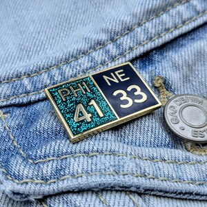 May include: A gold-colored pin with blue and green enamel. The pin has the text "PHI 41 NE 33" on it.
