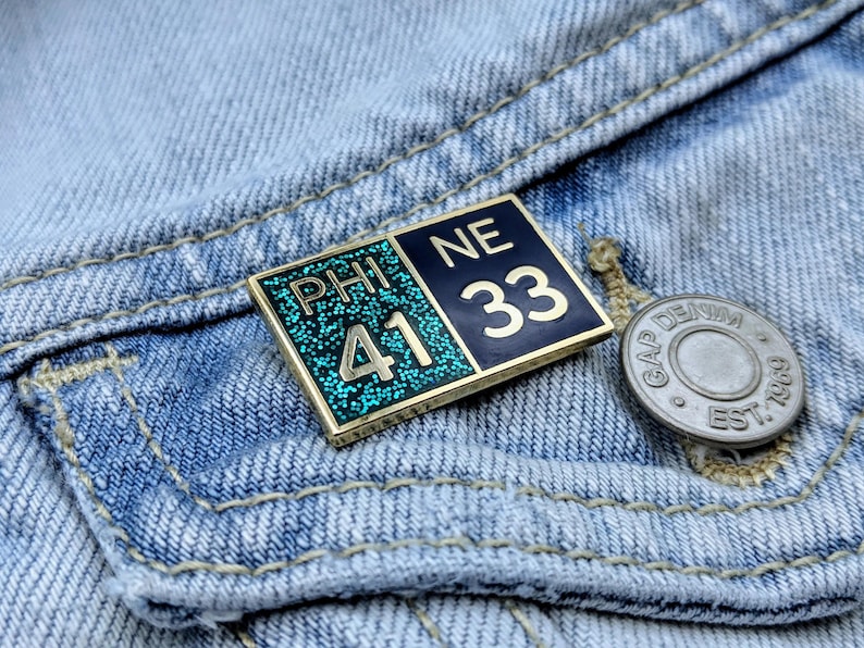 May include: A gold-colored pin with blue and green enamel. The pin has the text "PHI 41 NE 33" on it.