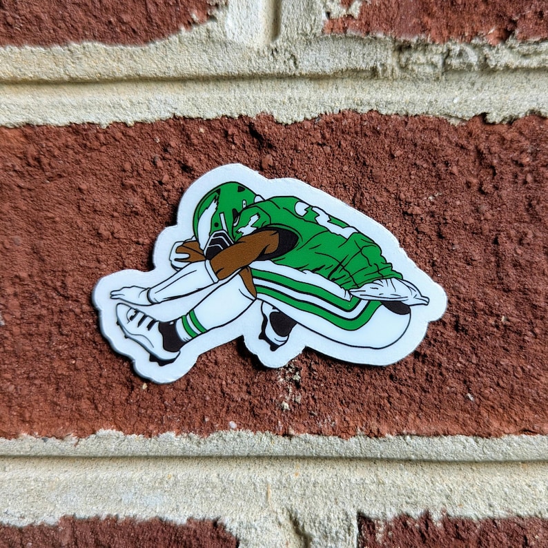 May include: A sticker featuring a football player in a green and white uniform, head down in a gesture of defeat. The sticker has a white border and is set against a red brick background.