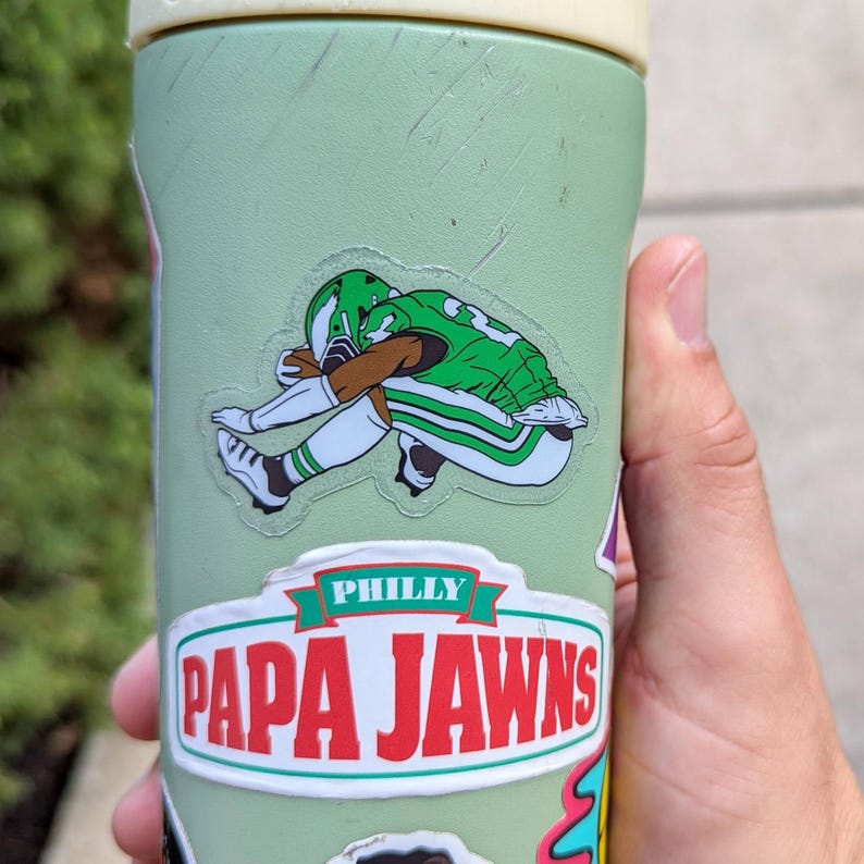 May include: A light green water bottle with a cream-colored lid. The bottle has a sticker of a football player in a green uniform with the number 33. Below the sticker is a sticker that says "PHILLY PAPA JAWNS" in red and white.
