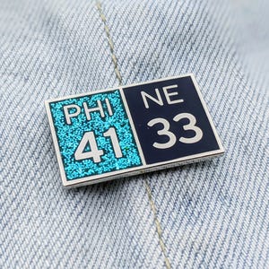May include: A blue and black enamel pin with the words "PHI 41" and "NE 33" on it.