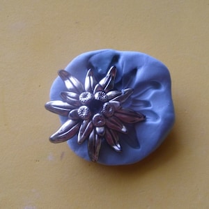 Puede incluir: Un amuleto de flor de edelweiss de metal plateado con cinco puntas y una superficie texturizada. El amuleto está sentado en un molde de silicona azul.