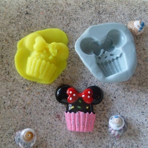 Peut inclure: Trois moules en silicone pour faire des cupcakes. Un moule est jaune et présente un cupcake avec un nœud sur le dessus. Un autre moule est gris et présente un cupcake avec un nœud sur le dessus. Le troisième moule est un cupcake en plastique avec un glaçage rose et blanc et un nœud noir sur le dessus.