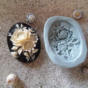 Puede incluir: Un camafeo blanco con un diseño floral, engastado en un marco ovalado negro. El camafeo está al lado de un molde de silicona del mismo diseño.