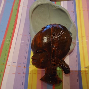 May include: A brown ceramic bust of a person with dark brown hair and a dark brown headband. The bust is sitting on a striped surface.