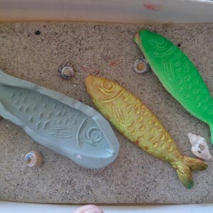 Puede incluir: Tres moldes con forma de pez, uno gris, uno amarillo y uno verde, sobre una superficie arenosa. Los moldes están detallados con escamas y aletas.