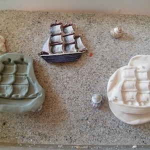 Puede incluir: Dos moldes de cerámica blanca de barcos de vela sobre una superficie arenosa de color marrón claro. Los moldes están detallados con líneas en relieve para crear el aspecto de las velas de un barco.