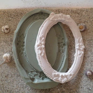 May include: A white oval picture frame with ornate detailing sits on a light brown surface. The frame is made of a smooth, white material and has a raised edge around the perimeter. The frame is slightly tilted to the right.