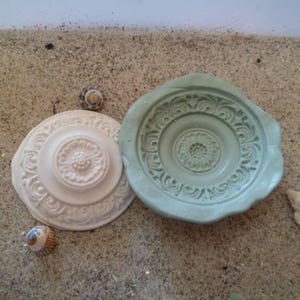 Op de afbeelding: Twee decoratieve ronde keramische borden met ingewikkelde ontwerpen. Het bord links is wit met een reliëfbloemmotief. Het bord rechts is groen met een reliëfbloemmotief.