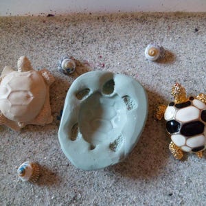 Puede incluir: Primer plano de un molde de silicona gris claro con forma de tortuga. El molde está sobre una superficie arenosa con varias conchas pequeñas blancas y azules. También hay una pequeña figurita de tortuga blanca y una figurita de tortuga más grande en blanco y negro con detalles dorados.