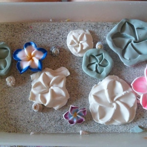 Puede incluir: Surtido de moldes de silicona en forma de flores blancas, rosas y azules sobre una superficie gris texturizada. Los moldes están diseñados para hacer jabón o velas decorativas.
