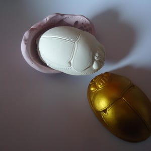 Peut inclure: Deux sculptures de scarabées, l'une blanche et l'autre dorée. Le scarabée blanc est dans un moule, tandis que le scarabée doré est sur une surface blanche.