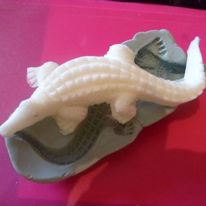 May include: A white, textured alligator figurine resting on top of a grey, flexible silicone mold. The alligator features detailed scales along its back and a long, curved tail, while the mold underneath shows the negative impression of the creature.