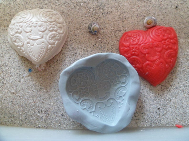 May include: Three heart-shaped moulds, one white, one red and one grey, on a sandy surface. The moulds have a decorative pattern.