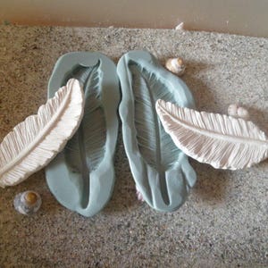 Puede incluir: Dos moldes de silicona para hacer decoraciones con forma de pluma. Los moldes son de color azul claro y tienen un diseño de pluma detallado. También se muestran dos decoraciones con forma de pluma blancas.