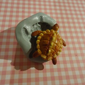 Puede incluir: Un molde de silicona gris para hacer un pequeño pastel con forma de tortuga u otro dulce comestible. El molde está vacío y listo para ser llenado con masa.