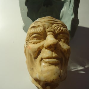 May include: A detailed, hand-crafted sculpture of a man's face. The sculpture is made of a light brown material and has a realistic expression. The man's face is wrinkled and has a slight smile.