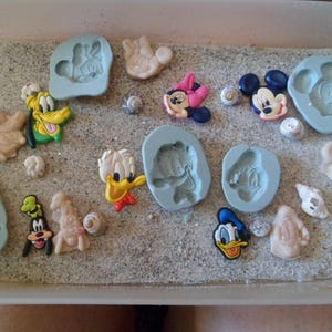 Puede incluir: Una colección de moldes de silicona con varios personajes de Disney, incluyendo Mickey Mouse, Minnie Mouse, Donald Duck y Goofy. Los moldes están dispuestos sobre una superficie gris claro con una textura similar a la arena.