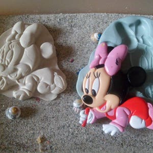 Peut inclure: Une figurine sculptée de Mickey Mouse de couleur claire et une figurine colorée de Minnie Mouse avec un nœud rose, une robe rouge et des gants roses. Un moule bleu est à côté de Minnie. Les figurines sont sur une surface texturée.