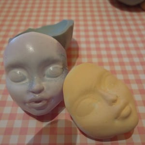 May include: Two unfinished doll heads, one with a light purple and blue face and the other with a light yellow face. Both heads have closed eyes and painted lips.