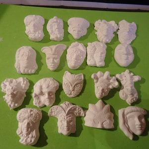 May include: A collection of 17 white plaster masks with various designs, including a cat, a jester, and a woman's face. The masks are arranged on a green surface.
