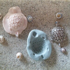 Puede incluir: Tres moldes con forma de tortuga sobre una superficie arenosa. Un molde es rosa y tiene un diseño de caparazón detallado. Otro molde es gris y tiene un patrón de panal. La tercera tortuga es plateada y tiene un diseño de caparazón detallado con piedras preciosas azules.