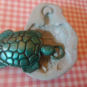 Puede incluir: Un dije de tortuga verde turquesa con una concha detallada y un anillo para sujetarlo a un collar o pulsera. El dije está sentado en un molde de silicona de una tortuga.