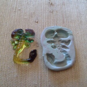 May include: A translucent scorpion-shaped craft element in shades of green, yellow, and brown, next to a light blue silicone mold. The scorpion has a curved tail and detailed body. The mold is in the shape of the scorpion.