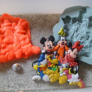 Könnte beinhalten: Ein Set Silikonformen zum Herstellen von Kuchendekorationen mit Disney-Charakteren. Die Formen sind blau und grau und zeigen Mickey Mouse, Minnie Mouse, Donald Duck, Goofy und Pluto. Die Formen werden neben einem Satz orangefarbener Kuchendekorationen gezeigt.