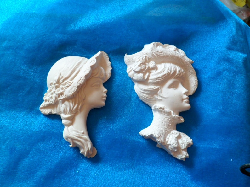 May include: Two white plaster wall decor pieces featuring the profile of a woman's face. Both women are wearing hats with intricate details.