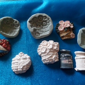 Puede incluir: Una colección de nueve moldes de cerámica blanca y gris para hacer manualidades en miniatura. Los moldes presentan diseños de una cesta de frutas, una maceta, una lata de leche y una cesta de flores.