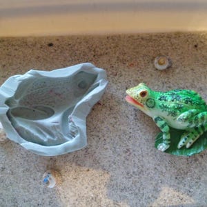 Könnte beinhalten: Eine grüne Froschfigur mit schwarzen Flecken, die auf einem grünen Blatt sitzt. Der Frosch ist aus Kunststoff und hat ein detailliertes Design. Der Frosch sitzt auf einer weißen Oberfläche.