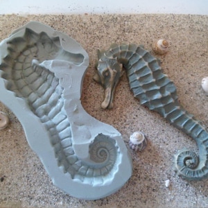 Puede incluir: Un molde de silicona gris de un caballito de mar, con una superficie texturizada detallada. El molde está sobre una superficie arenosa con pequeñas conchas.