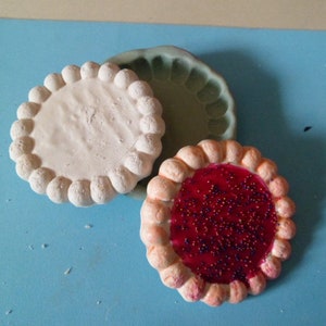 Peut inclure: Deux croûtes à tarte en céramique blanche et une remplie de gelée rouge et de pépites violettes. Les croûtes à tarte sont sur une surface bleue.