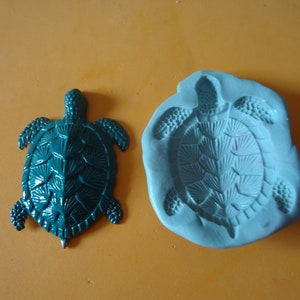 Puede incluir: Un molde de tortuga verde azulado y una figurita de tortuga verde azulado. El molde está hecho de silicona y tiene un diseño detallado. La figurita está hecha de plástico y tiene un acabado liso.