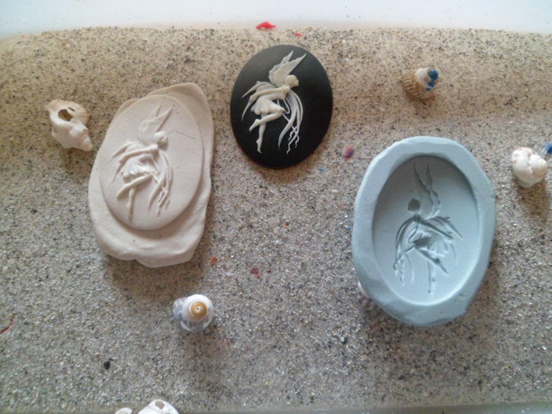 Puede incluir: Un surtido de materiales de manualidades con tem&aacute;tica de hadas. Incluye un camafeo ovalado negro con un dise&ntilde;o de hada blanca, un molde ovalado gris claro y un relieve de arcilla beige claro. Peque&ntilde;as conchas marinas dispersas.