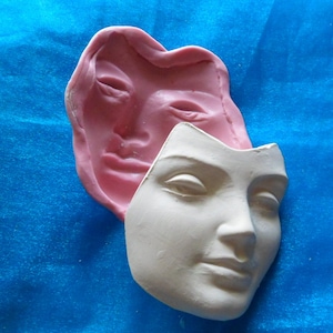 Peut inclure: Un masque facial en plâtre blanc avec un moule en silicone rose derrière. Le moule est une empreinte négative du masque facial.