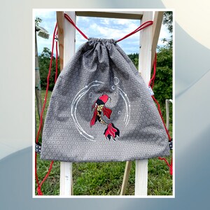 Puede incluir: Mochila gris con cordón y estampado geométrico, con diseño de pez koi rojo y dorado. La mochila tiene cordones rojos y se exhibe sobre una estructura de madera. El fondo muestra un campo verde y un cielo azul.