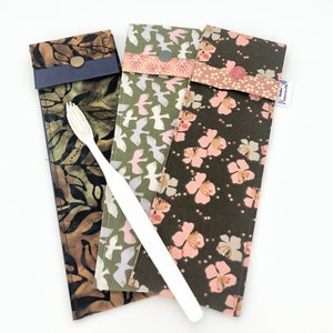 Peut inclure: Trois étuis à brosse à dents en tissu avec différents motifs : un batik marron, un imprimé d'oiseaux verts et un motif floral vert foncé. Une brosse à dents blanche est posée sur les étuis. Chaque étui a une fermeture à bouton.