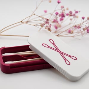 May include: A rectangular storage case with a deep magenta base and a white lid featuring a magenta graphic of two crossed cotton swabs. Inside the open case are two wooden cotton swabs with white tips, positioned horizontally within the molded slots.