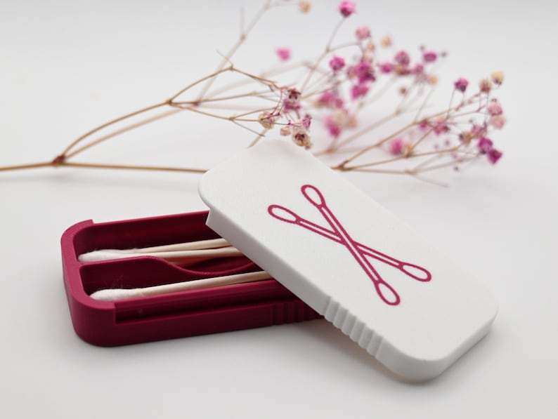 May include: A rectangular storage case with a deep magenta base and a white lid featuring a magenta graphic of two crossed cotton swabs. Inside the open case are two wooden cotton swabs with white tips, positioned horizontally within the molded slots.