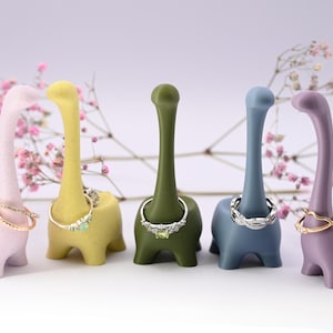 May include: Five colorful dinosaur-shaped ring holders. Each holder is a different color: pink, yellow, green, blue, and purple. Each holder has a ring on it.