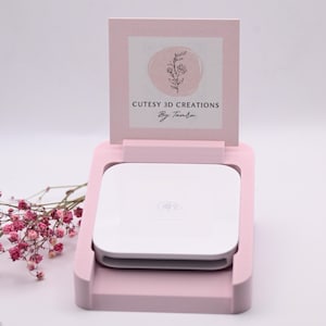 May include: A light pink 3D-printed plastic stand holding a white square payment card reader. The stand features an upright back panel displaying a logo with a pink circle, a floral line drawing, and the text CUTESY 3D CREATIONS By Tamka.