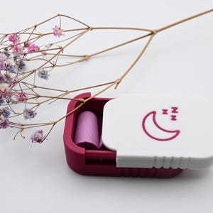 May include: A small, burgundy case with a white lid featuring a crescent moon and sleeping symbols, containing two purple earplugs. A sprig of dried flowers with pink and white blossoms rests nearby. The case is open, revealing the earplugs.