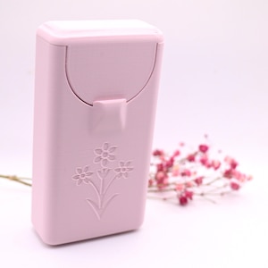 May include: A rectangular light pink plastic case with a hinged lid and a latch mechanism. The front surface features an embossed design of three stylized flowers with stems and leaves. The case has a smooth, matte finish and rounded corners.